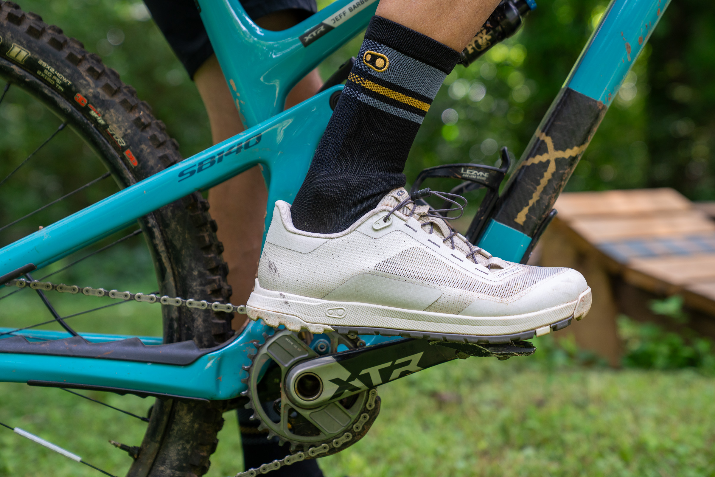 A close-up view of a cyclist's foot on a mountain bike pedal, showcasing a white cycling shoe with a textured surface and gray laces. The cyclist is wearing a black sock with gray and gold stripes. The bike features a teal frame and a visible gear mechanism labeled "XTR" on the crankset, with a blurred background of greenery.