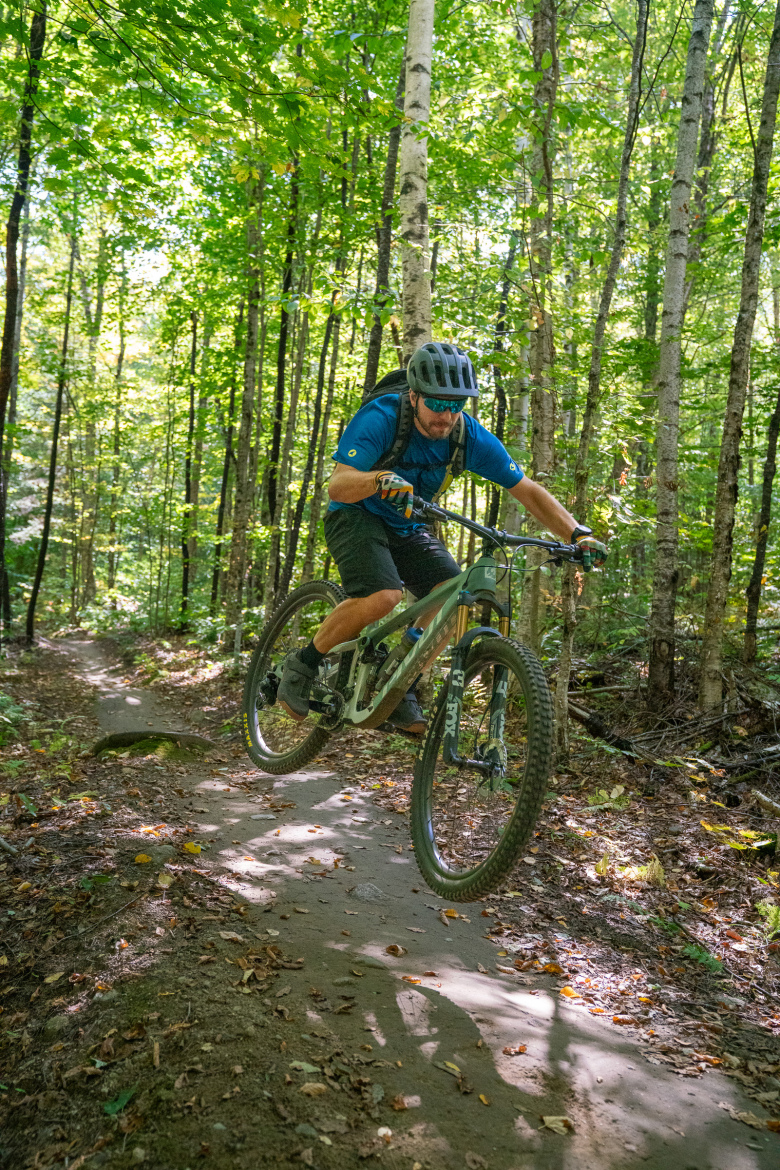 A mountain biker performing a jump on a dirt trail surrounded by lush green trees in a forest setting. The rider is wearing a helmet, sunglasses, and a blue jersey with black shorts, showcasing an active and adventurous moment.