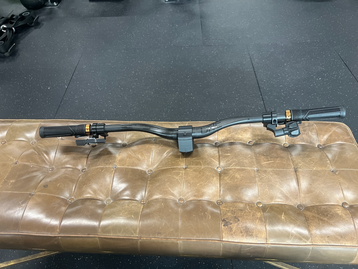 A close-up view of a black bicycle handlebar resting on a tufted brown leather bench in a gym setting. The handlebar features textured grips and a gear shifter, showcasing its functional design against the backdrop of a gym environment.