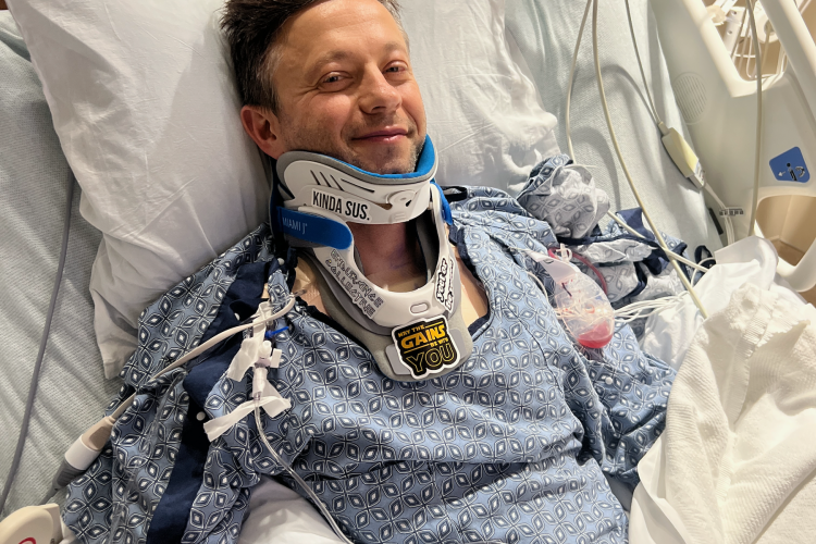 A man in a hospital bed wearing a blue patterned gown, a neck brace, and an IV in his arm. He has a cheerful expression and a sticker on his neck brace that reads "Kinda Sus." Medical equipment is visible in the background.