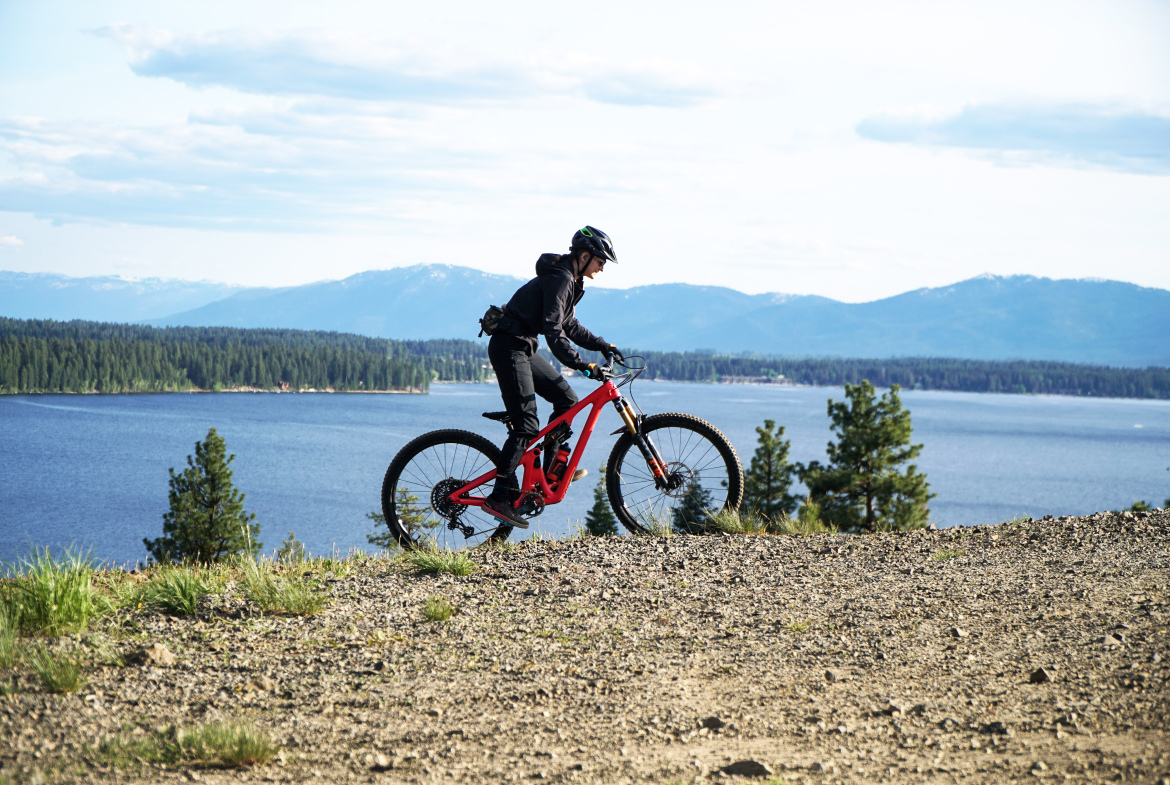 A cyclist riding a red mountain bike on a gravel trail overlooking a lake and mountain landscape. The rider is wearing a helmet and has a black jacket and pants, with trees and hills visible in the background under a partly cloudy sky.