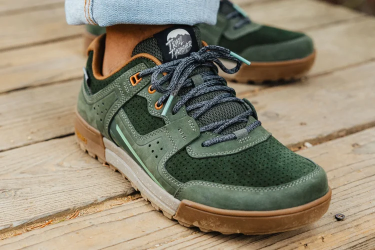 Close-up of a pair of green sneakers with a mix of suede and mesh materials, featuring grey laces and orange accents. The shoes are positioned on a wooden surface, showcasing their unique design details and textures.