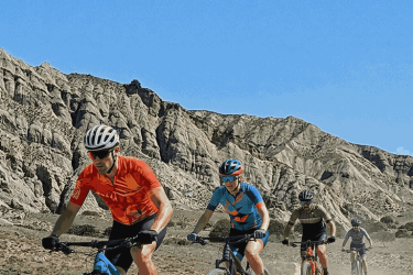 A group of five mountain bikers riding on a dirt trail through a rugged landscape with rocky formations in the background. The cyclists are dressed in colorful cycling gear, and dust is being kicked up as they pedal through the terrain under a clear blue sky. Vashlovani National Park Adventure Loop mountain bike trail.