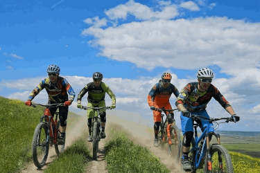 Four mountain bikers riding on a dusty trail surrounded by green grass under a bright blue sky with scattered clouds. Vashlovani National Park Adventure Loop mountain bike trail.