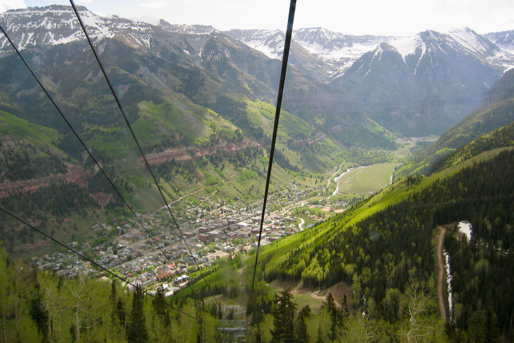 A scenic view from a cable car, showcasing a lush green valley surrounded by mountains. The image features a small town nestled in the valley, with winding roads and patches of snow on nearby peaks. Cable lines from the gondola can be seen stretching across the foreground, adding a sense of depth to the landscape.