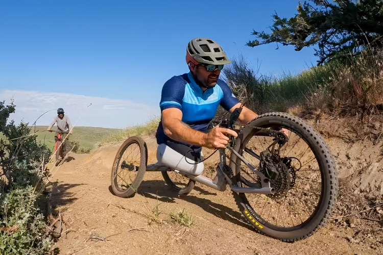 A person riding a specialized mountain bike with three wheels on a dirt trail, navigating a downhill slope. The rider is wearing a blue and black cycling jersey, a helmet, and sunglasses. In the background, another individual is visible riding a traditional bicycle on a parallel path. The scene is surrounded by greenery under a clear blue sky.