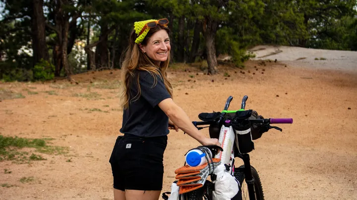 A woman with long hair smiles while standing next to a bicycle, wearing a black shirt and shorts. She has a bright yellow headband and her bike is equipped with colorful accessories. The background features a dirt path and trees, suggesting a natural outdoor setting.
