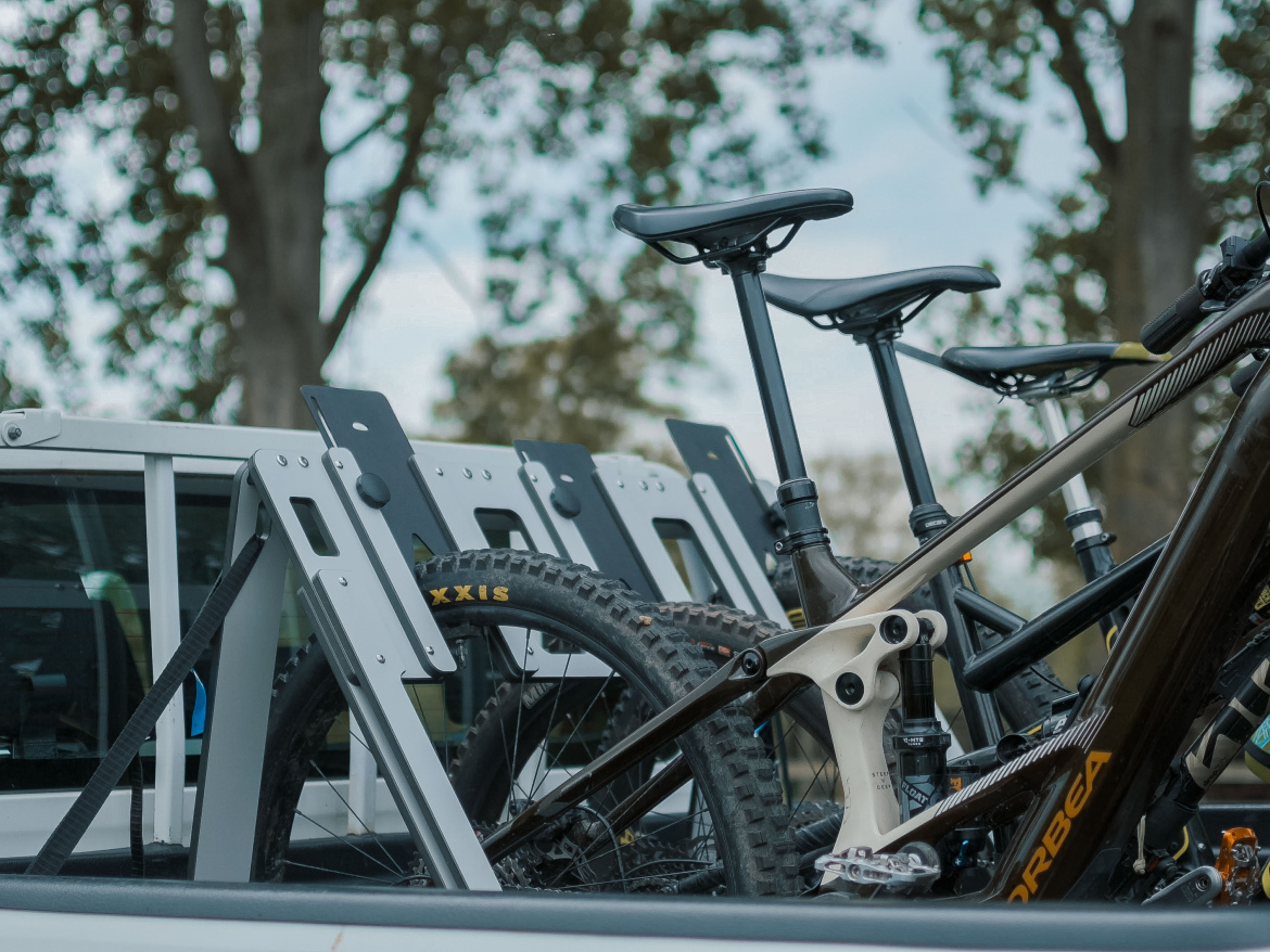 A close-up view of several mountain bikes secured in the bed of a pickup truck, with bike racks visible in the background and trees in the distance.