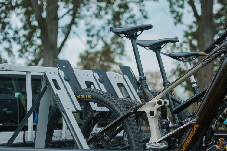 A close-up view of several mountain bikes secured in the bed of a pickup truck, with bike racks visible in the background and trees in the distance.