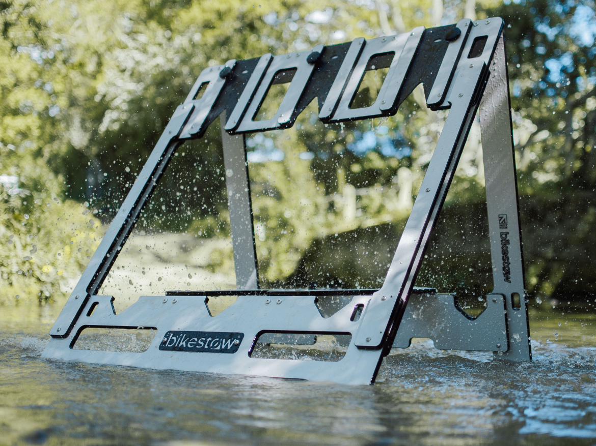 A bike storage stand partially submerged in water, creating splashes around it. The stand features a modern design with a white and black color scheme and is labeled with the brand name. Lush greenery can be seen in the background, indicating an outdoor setting.