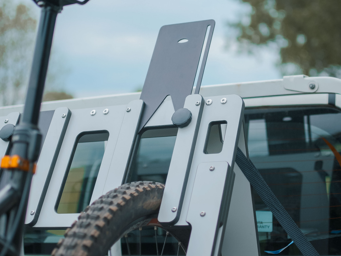 A close-up view of a bike rack mounted on the rear of a vehicle, featuring a metallic structure with a vertical plate for securing a bicycle. The rack includes a wheel holder with a textured tire visible and a black strap securing the bike in place. In the background, the vehicle's rear window is partially visible, with trees and a cloudy sky.