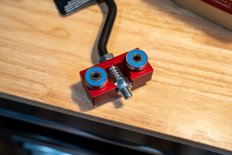 Close-up view of a red aluminum block with two blue bearings and a threaded bolt in the center. The component is mounted on a wooden surface, and a black hose connects to one side.