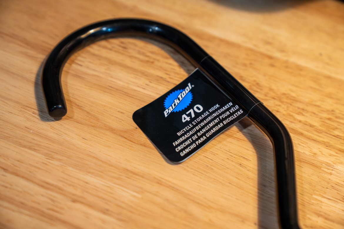 A black bicycle storage hook, labeled "Park Tool 470," resting on a wooden surface. The tag provides information in multiple languages about its use for hanging bicycles.