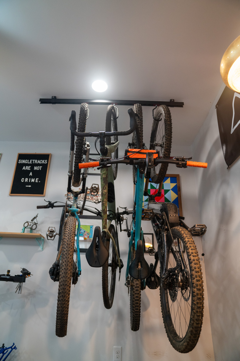 Four bicycles are hung vertically from a ceiling-mounted rack in a room. The bicycles display a variety of frame colors, including a camo pattern and bright blue. The space features a light fixture above and a wall decorated with a sign that reads "SINGLETRACKS ARE NOT A CRIME." Several decorative items, including a colorful artwork and tools, are visible on nearby shelves.