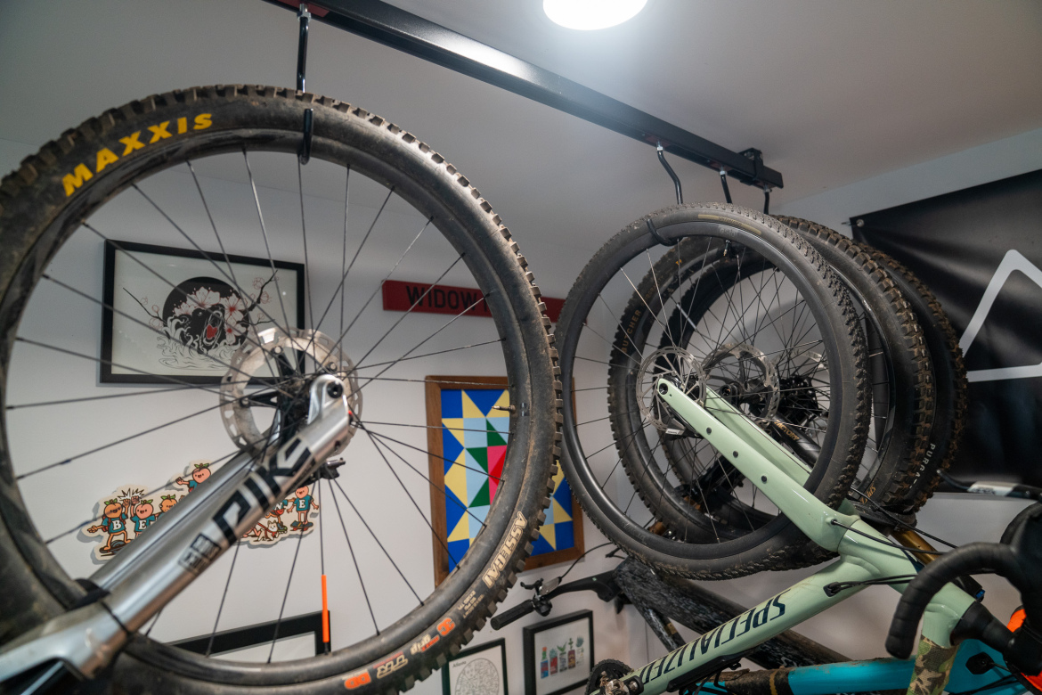 A collection of bicycle wheels suspended from a ceiling rack in a garage. The wheels feature various tire brands and patterns. In the background, there are framed artworks and decorations on the walls, including a colorful geometric design and a sign labeled "WIDOW." The scene is well-lit with a ceiling light overhead.