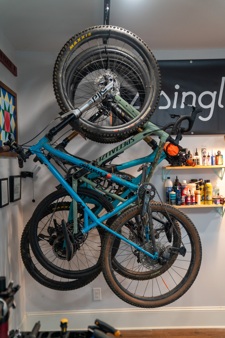 Alt text: "Two mountain bikes hanging on a wall-mounted rack, one blue and one green, with tools and bike maintenance supplies visible on a shelf in the background."