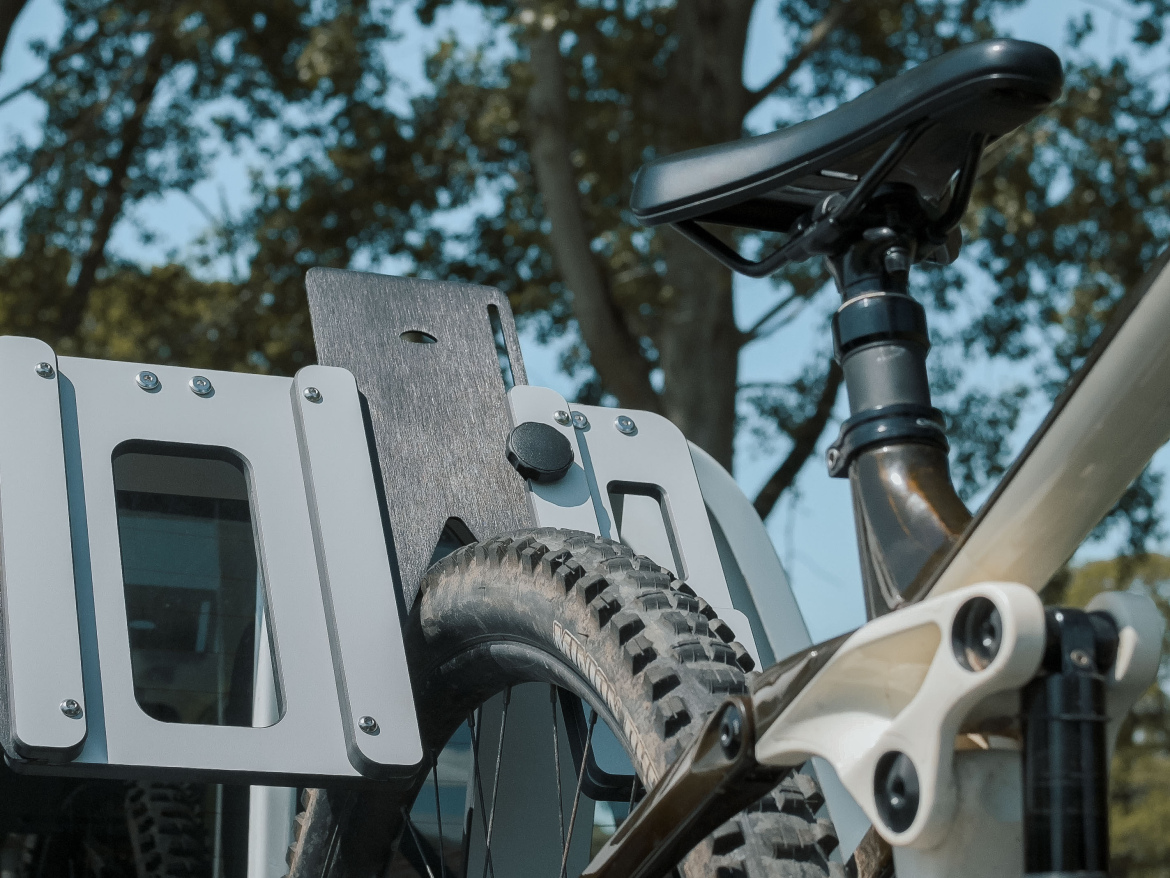 A close-up view of a bicycle's rear wheel and saddle, showcasing a modern bike rack with a sturdy mount. The background features blurred greenery, indicating an outdoor setting.