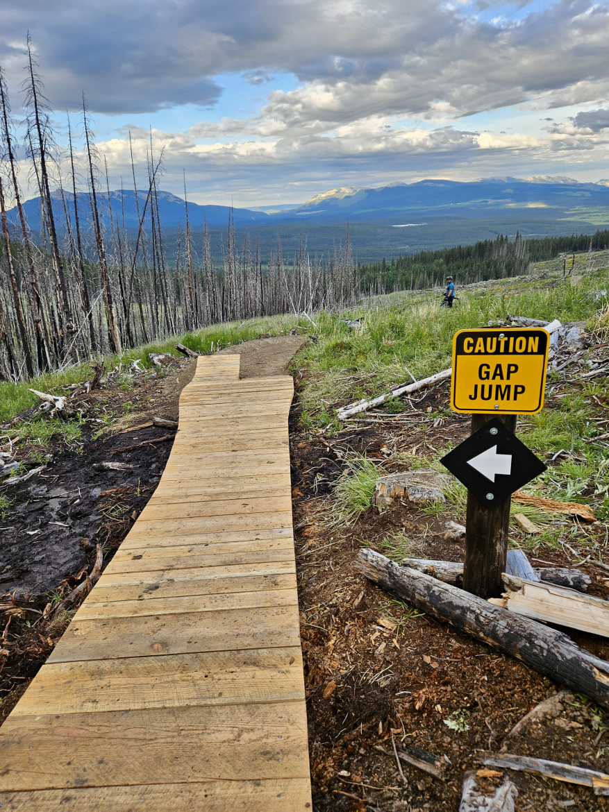 A wooden pathway winds through a mountainous landscape, leading downhill amid charred tree trunks and lush greenery. In the foreground, a caution sign indicating a "Gap Jump" is visible, with an arrow pointing to the left. The background features a scenic view of mountains and valleys under a partly cloudy sky. A person can be seen in the distance, walking along the trail.