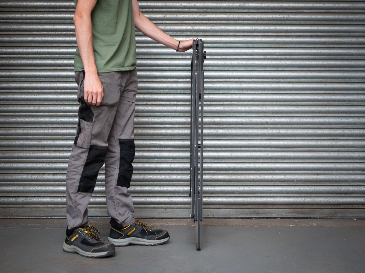A person standing next to a closed, metallic roll-up door, holding a folded portable table with one hand. They are wearing a green shirt, gray cargo pants with black patches, and sturdy footwear. The background features a solid gray floor and a slightly reflective surface.