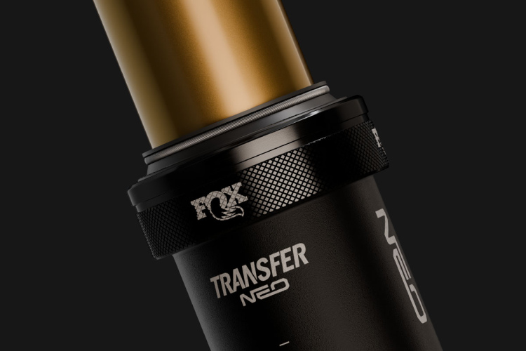 Close-up image of a black and gold bicycle component, featuring a cylindrical shaft with a shiny gold finish and a textured black adjustment knob. The product is labeled with the name "TRANSFER NEO" and the logo is visible, set against a dark background.