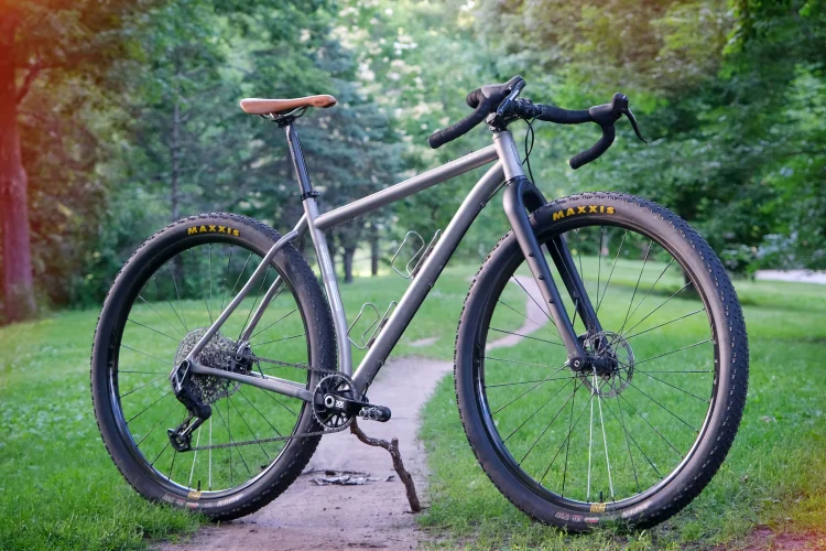 A rugged mountain bike with a sleek silver frame and brown leather saddle, parked on a dirt path surrounded by green grass and trees in a natural outdoor setting. The bike features wide tires with Maxxis branding and a minimalist design, displaying a strong and adventurous look.