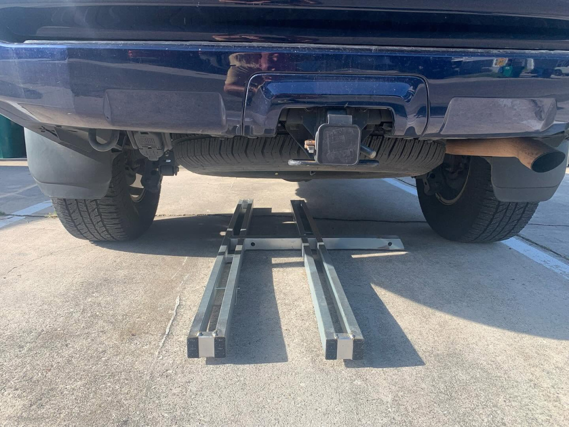 A close-up view of the rear of a blue vehicle, showing the tow hitch and exhaust pipe. Below the vehicle, there are two parallel metal tracks positioned on the ground, indicating a setup for loading or towing. The background features a paved surface.