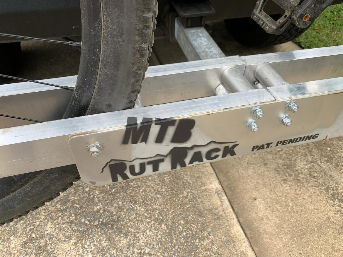 An aluminum bicycle rack designed for mountain bikes, prominently displaying the text "MTB Rut Rack" along with the phrase "PAT. PENDING." The rack is shown mounted to a vehicle, holding a mountain bike tire in place, against a background of gravel and grass.