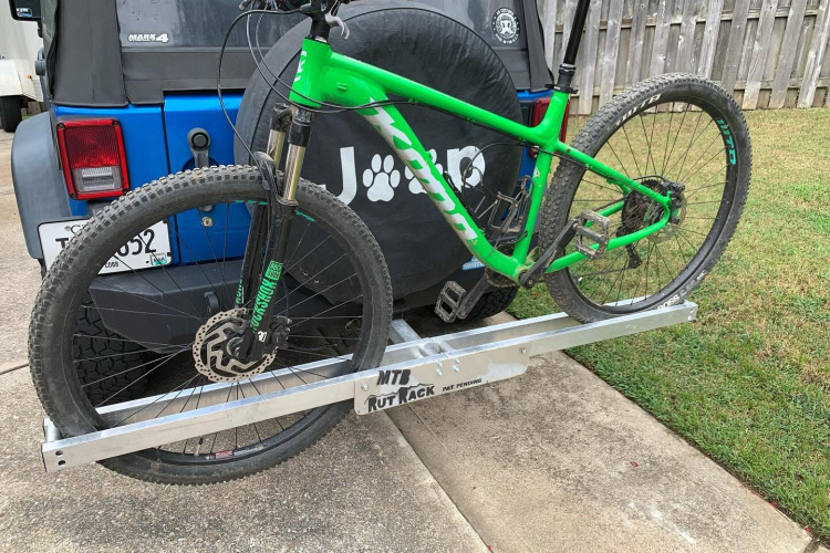 A green mountain bike mounted on a silver hitch rack attached to a blue Jeep parked on a driveway. The bike features thick tires, disk brakes, and a prominent suspension fork. In the background, a wooden fence and grassy area are visible.