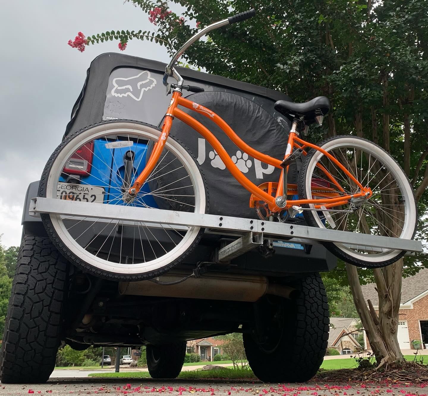 An orange bicycle is mounted on the back of a Jeep, with a visible spare tire cover featuring the word "Jeep." The scene is set against a cloudy sky, surrounded by greenery, and colorful petals scattered on the ground. The perspective is from below the vehicle, highlighting the bicycle and tire.