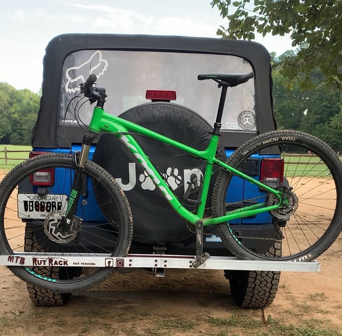 A bright green mountain bike secured to a bike rack on the back of a blue Jeep. The bike features black tires with visible tread and is positioned next to a spare tire cover with a paw print design. The background includes a dirt path and green trees, suggesting an outdoor setting.