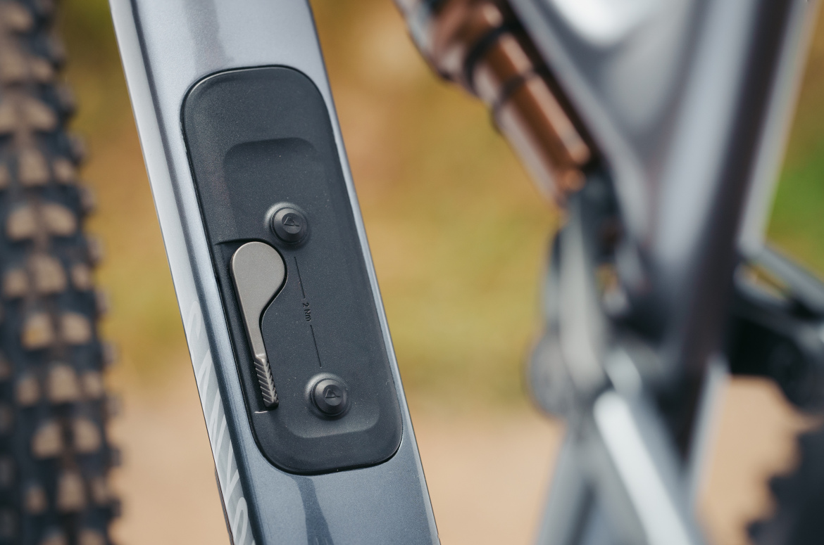 Close-up of a bike frame showing a mechanical component with a textured lever and adjustment markings. The background is blurred, highlighting the details of the component.