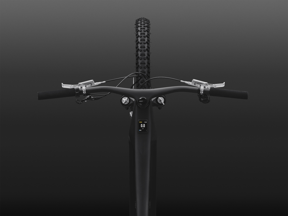 A top-down view of a mountain bike's handlebars against a dark background. The bike features a sleek design with black grips, brake levers, and a compact display screen showing speed metrics. The front wheel, partially visible, has a rugged tire tread suitable for off-road terrain.