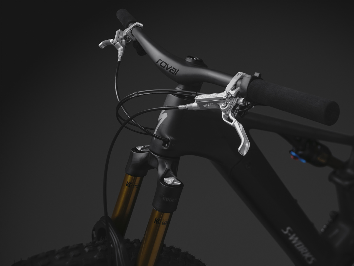 Close-up view of a high-performance mountain bike's handlebars and suspension forks, featuring sleek black handlebars with rubber grips, brake levers, and visible brake cables. The bike's fork legs are gold, highlighting its premium construction, against a dark background that emphasizes the bike's modern design and quality components.