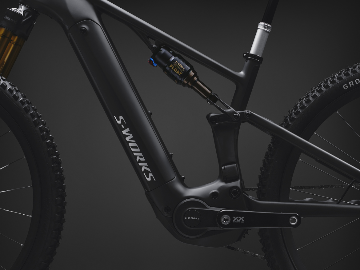Close-up view of a black mountain bike frame featuring the "S-WORKS" logo, showcasing the battery compartment and rear suspension components. The bike has rugged, knobby tires and a sleek, modern design against a dark background.