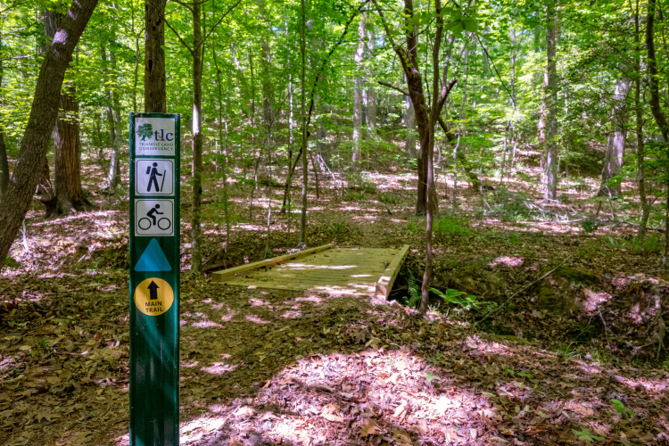 A trail sign indicating access for hikers and cyclists, with a wooden bridge in the background surrounded by lush green trees and fallen leaves on the ground. The sign features symbols for walking and biking, along with directional arrows leading to the main trail.