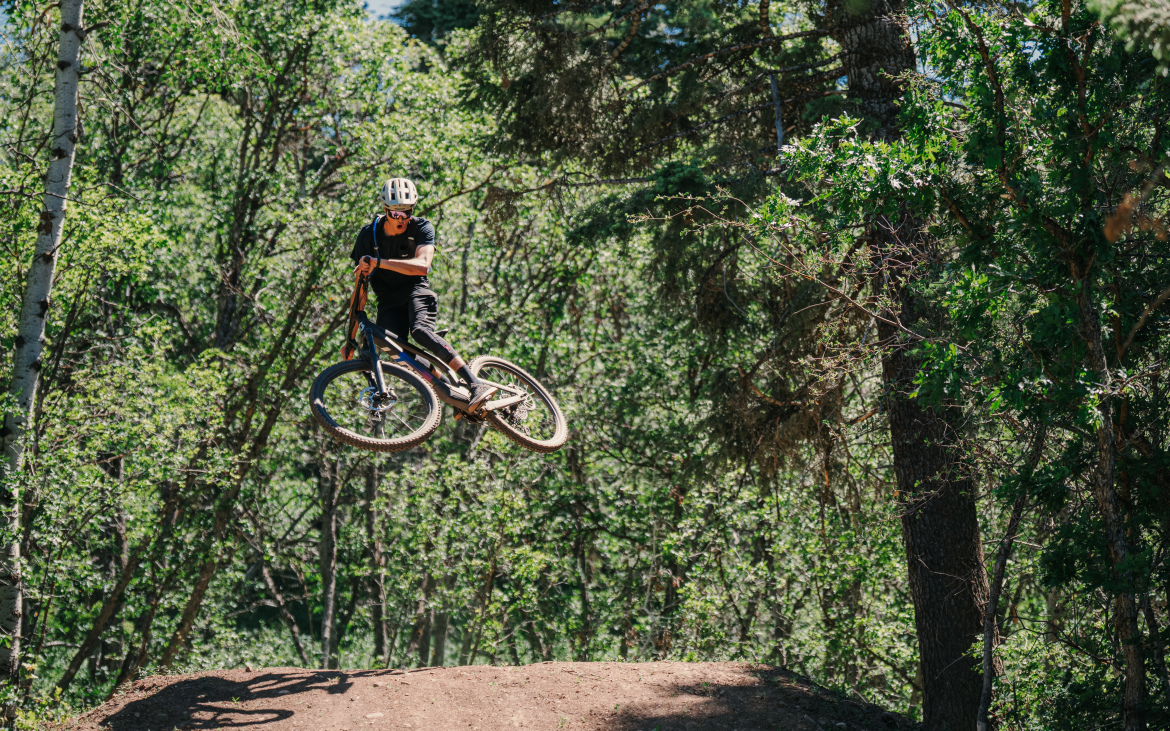 A mountain biker performing a jump over a dirt ramp in a forested area, surrounded by trees and greenery. The biker is wearing a helmet and is captured mid-air, showcasing a dynamic and thrilling moment of the sport.