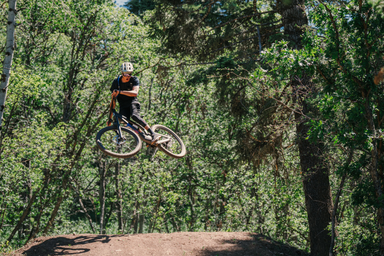 A mountain biker performing a jump over a dirt ramp in a forested area, surrounded by trees and greenery. The biker is wearing a helmet and is captured mid-air, showcasing a dynamic and thrilling moment of the sport.