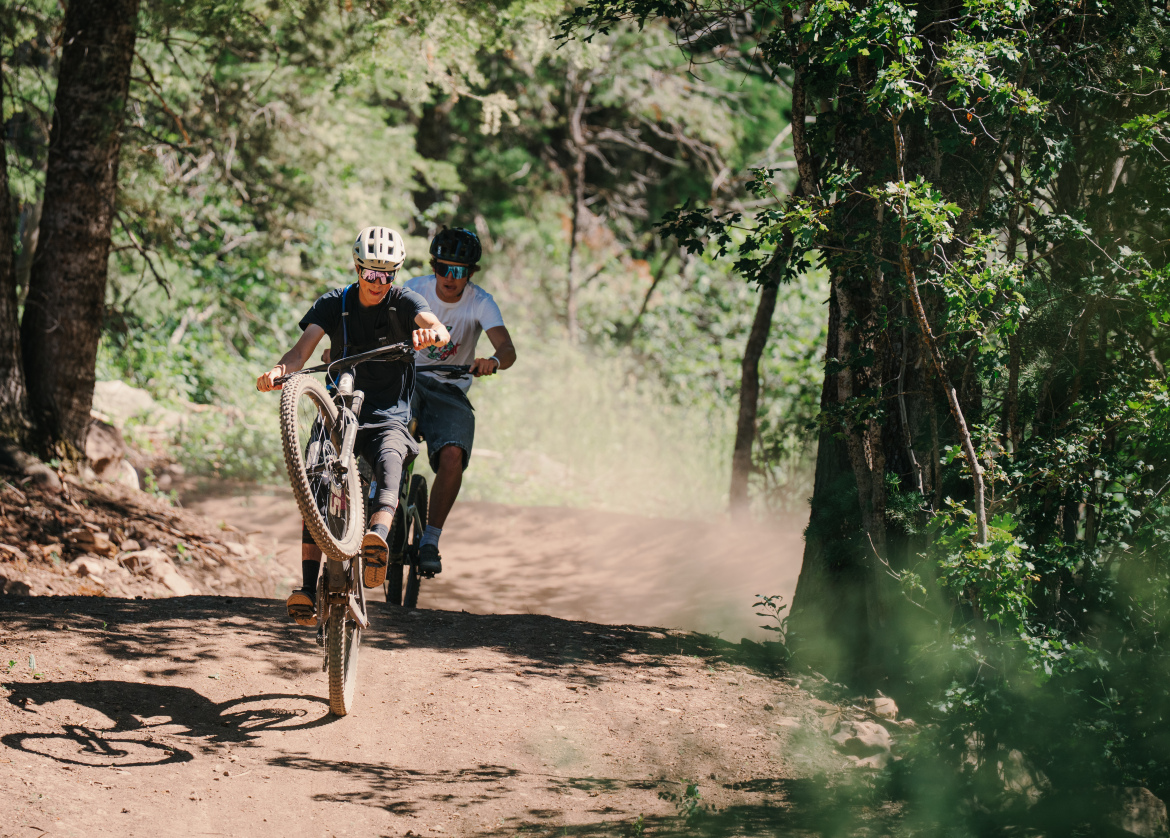 Two young men are riding mountain bikes on a dirt trail surrounded by trees. One rider is performing a wheelie, lifting the front wheel off the ground, while the other follows closely behind. Sunlight filters through the leaves, creating a vibrant, lively atmosphere as dust is kicked up from the trail.