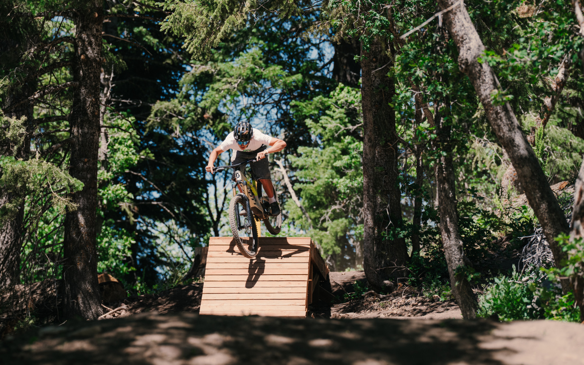 A mountain biker jumps off a wooden ramp in a forested area, surrounded by tall trees and bright green foliage, capturing a moment of action and adventure in the outdoors.