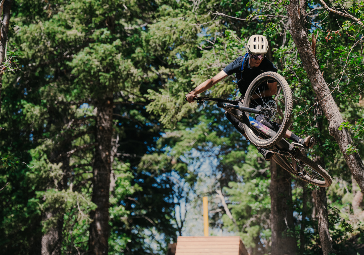A mountain biker performing a jump over a wooden ramp in a forested area, with trees in the background and bright sunlight illuminating the scene. The rider is wearing a helmet and protective gear.