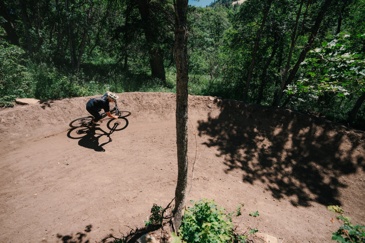 A mountain biker leans into a turns on a dirt track surrounded by lush greenery, with a tree positioned near the center of the bend. The bike kicks up dust as the rider navigates the course under bright sunlight.