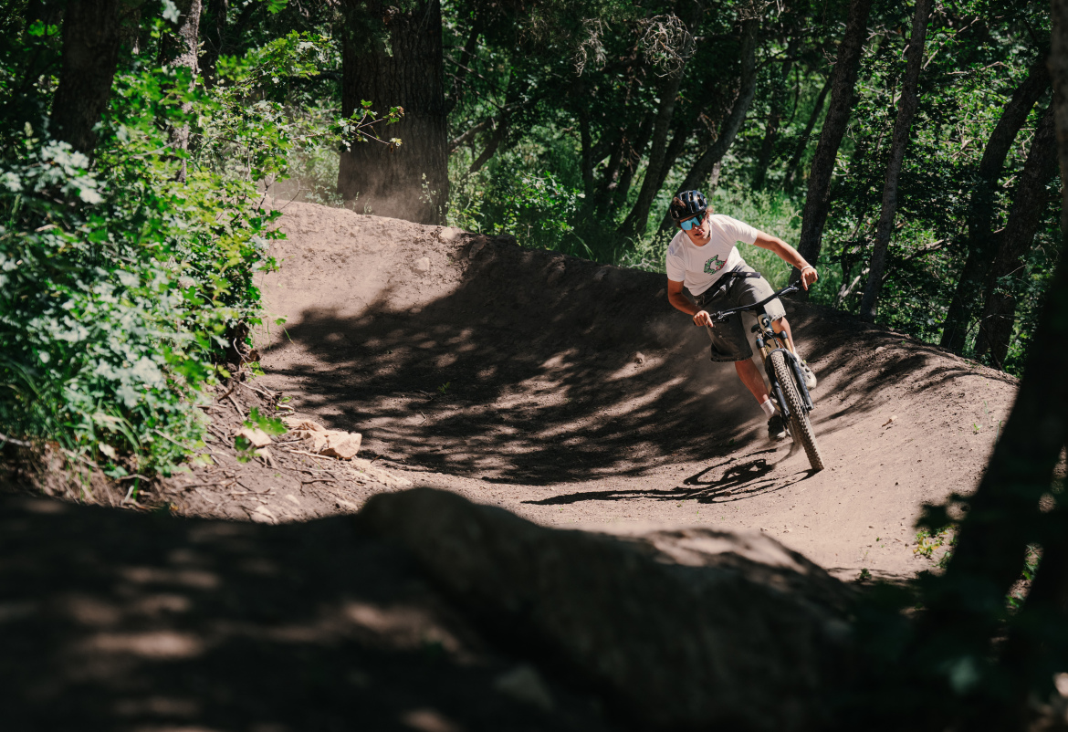 A mountain biker navigates a dirt path in a forested area, leaning into a turn with dust kicked up behind him. The scene is surrounded by trees and greenery, capturing the essence of outdoor biking activity.