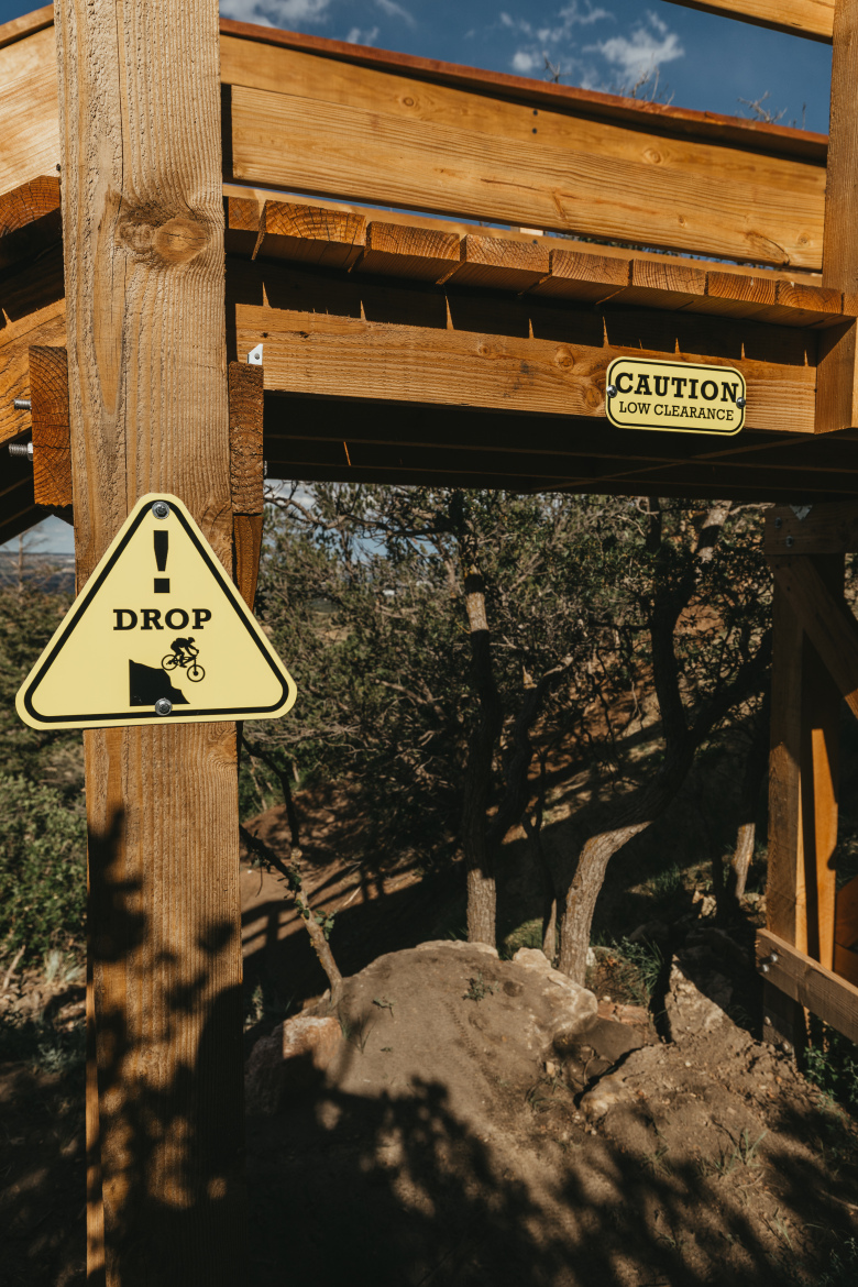 A wooden structure with two warning signs, one indicating a "DROP" for cyclists and the other cautioning about "LOW CLEARANCE." The background features trees and a dirt slope under a clear blue sky.