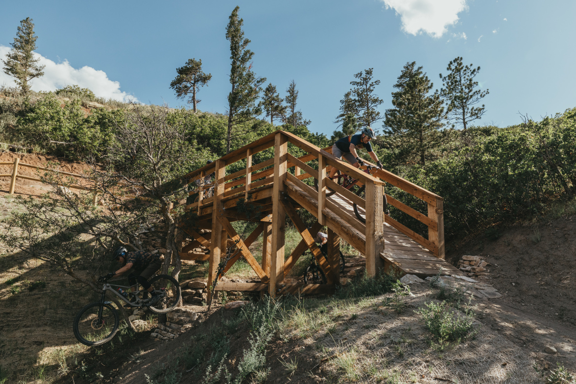 A mountain biker descends a wooden bridge in a forested area, with green vegetation and scattered trees surrounding the scene. The biker is focused and wearing a helmet, while another bike is positioned on the ground near the base of the bridge. The landscape features a mix of sunlight and shadows, creating an outdoor adventure atmosphere.