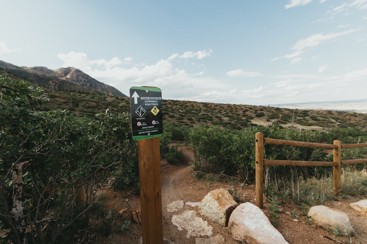 A trail sign indicating the entrance to the Neurotoxin Downhill Bike Trail, positioned beside a dirt path with surrounding greenery and a mountainous landscape in the background under a partly cloudy sky.