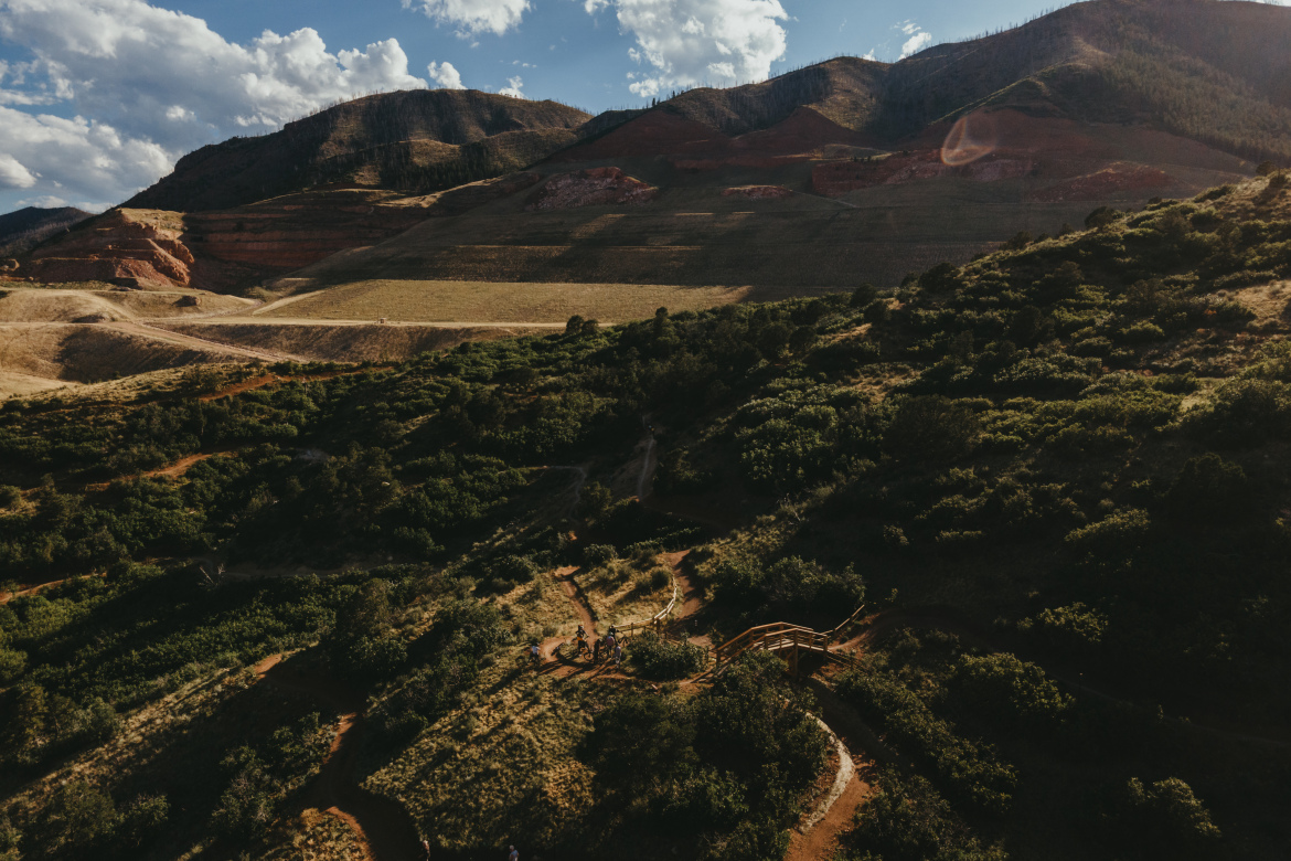 Aerial view of a hilly landscape featuring a mix of green vegetation and dry terrain, with rust-colored hills in the background. A wooden path winds through the lush areas, leading to a small structure. The sky is partially cloudy, with sunlight casting shadows across the landscape.