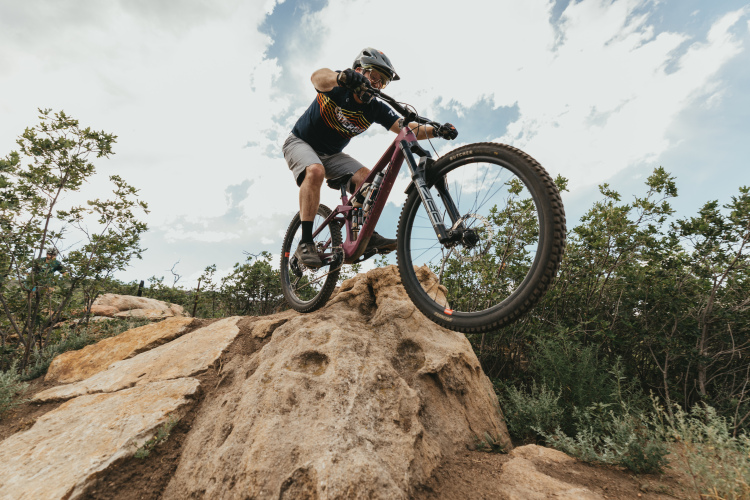 A mountain biker performing a jump over a large rock formation, with green shrubs and trees in the background under a partly cloudy sky. The rider is wearing a helmet and protective gear, demonstrating an action-packed moment in an outdoor setting.