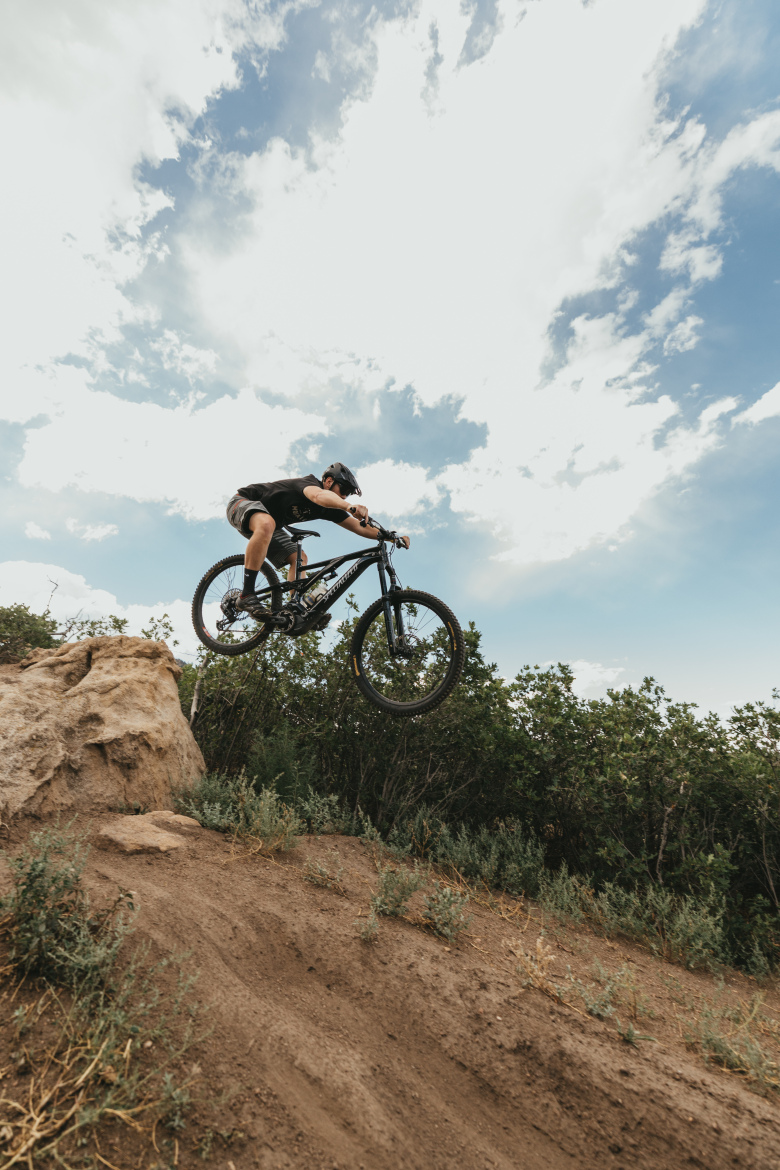 A mountain biker performs a jump off a rocky ledge, soaring through the air against a backdrop of blue sky with scattered clouds. The cyclist, wearing a helmet and casual riding gear, appears focused and in control as they lift their bike above a dirt trail surrounded by greenery.