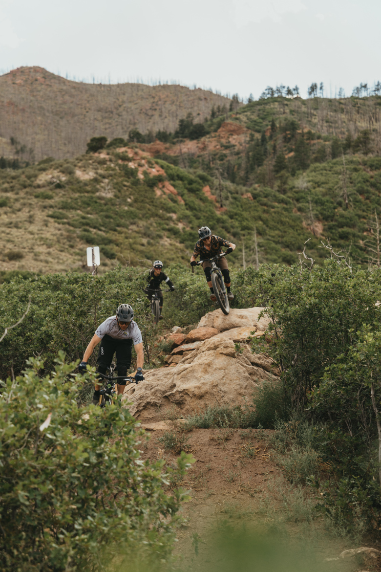 Three mountain bikers are navigating a rocky trail surrounded by lush greenery and hills. One rider is descending from a rock formation, while the others are further back on the trail, showcasing action and excitement in a natural landscape. The scene is set on an overcast day, adding a dramatic backdrop to the outdoor adventure.