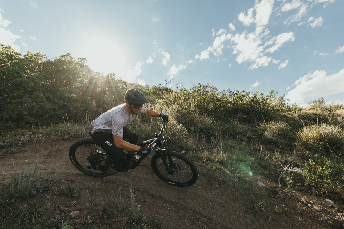 A cyclist in a helmet and riding gear skillfully navigates a dirt trail surrounded by greenery, with the sun shining in the background against a partly cloudy sky.